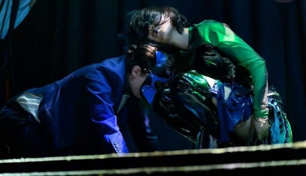 Two costumed performers in green and blue embracing under stage light.