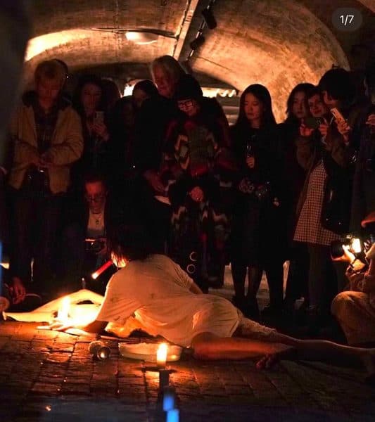 A candlelit crypt with onlookers gathered around a reclining performer.