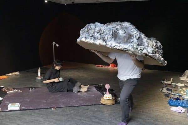 A performer carrying a large foil-wrapped boulder across a studio floor.