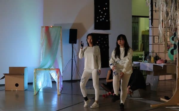 Two performers in white moving through a softly lit gallery.