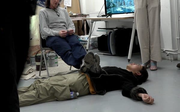 A performer lying on the floor as a seated visitor rests their feet on them.